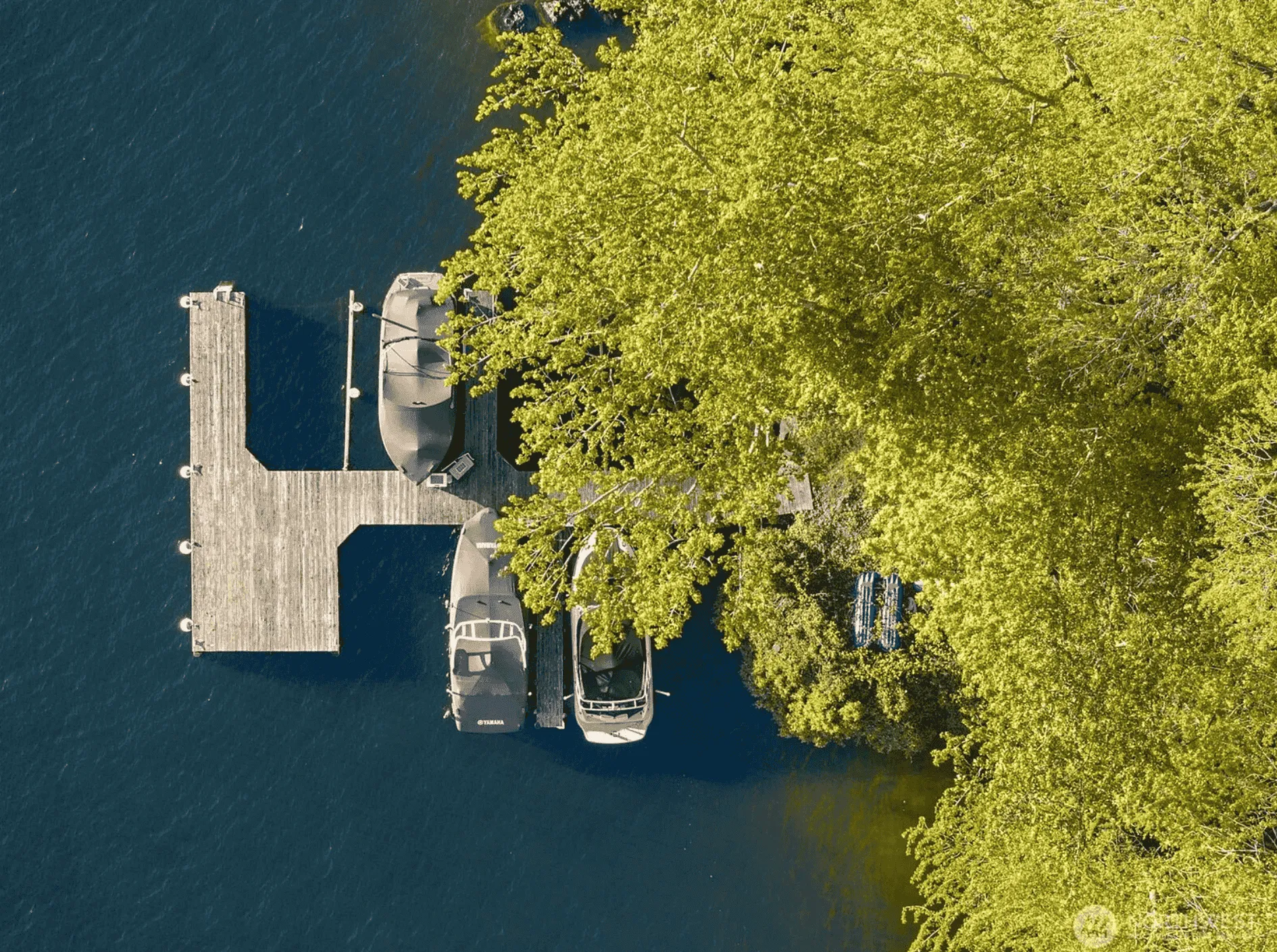 Overhead aerial view of a private wooden dock extending into deep blue water beside a tree-lined shoreline, with two boats moored alongside.